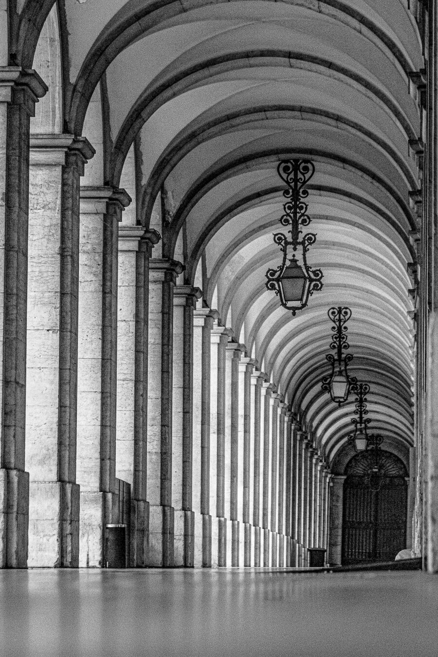 Black‑and‑white photograph of a long stone arcade in Lisbon, Portugal, captured from a low floor‑level perspective. Repeating arches and columns recede into the distance, creating a rhythmic pattern of light and shadow. Ornate wrought‑iron lanterns hang from the vaulted ceiling, leading the eye toward a dark arched gate at the far end. The image emphasizes symmetry, texture, and the quiet grandeur of classical architecture.
