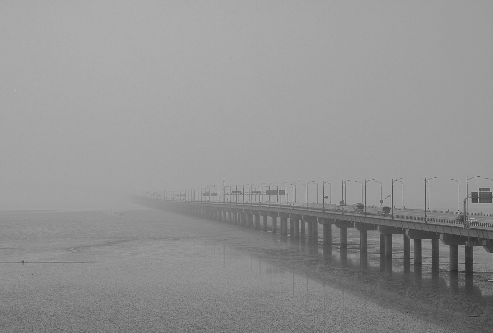 Incheon Bridge in South Korea stretches across calm tidal flats under a dense early‑morning fog. The long span of concrete pillars and streetlights fades gradually into the mist, with a few cars visible on the roadway. The monochrome tones emphasize the quiet, atmospheric mood, giving the scene a sense of distance and stillness.