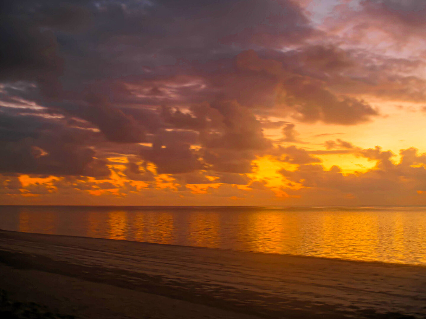 Dawn over the Indian Ocean from Mombasa, Kenya, captured in 2016. The horizon glows with deep amber and gold as sunlight breaks through scattered clouds, reflecting across calm water. The beach in the foreground lies in soft shadow, while the sky transitions from dark purple and grey to vivid orange, creating a serene and luminous start to the day.