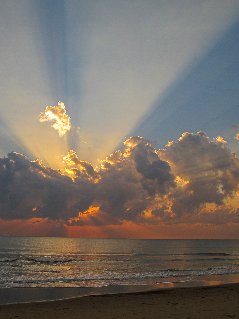Dawn breaking over the Gulf of Thailand from Aleenta, Pranburi, Thailand. Golden sunlight streams through dramatic clouds, casting radiant beams across the sky and shimmering reflections on calm sea waves, with the beach in soft shadow below.