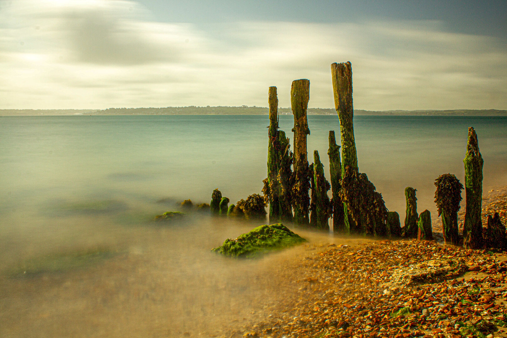 Long exposure of old wooden groyne at Lepe, Hampshire, England. Weathered posts covered in green algae stand in calm, misty water under a soft, cloudy sky, with pebbles and moss along the shoreline.