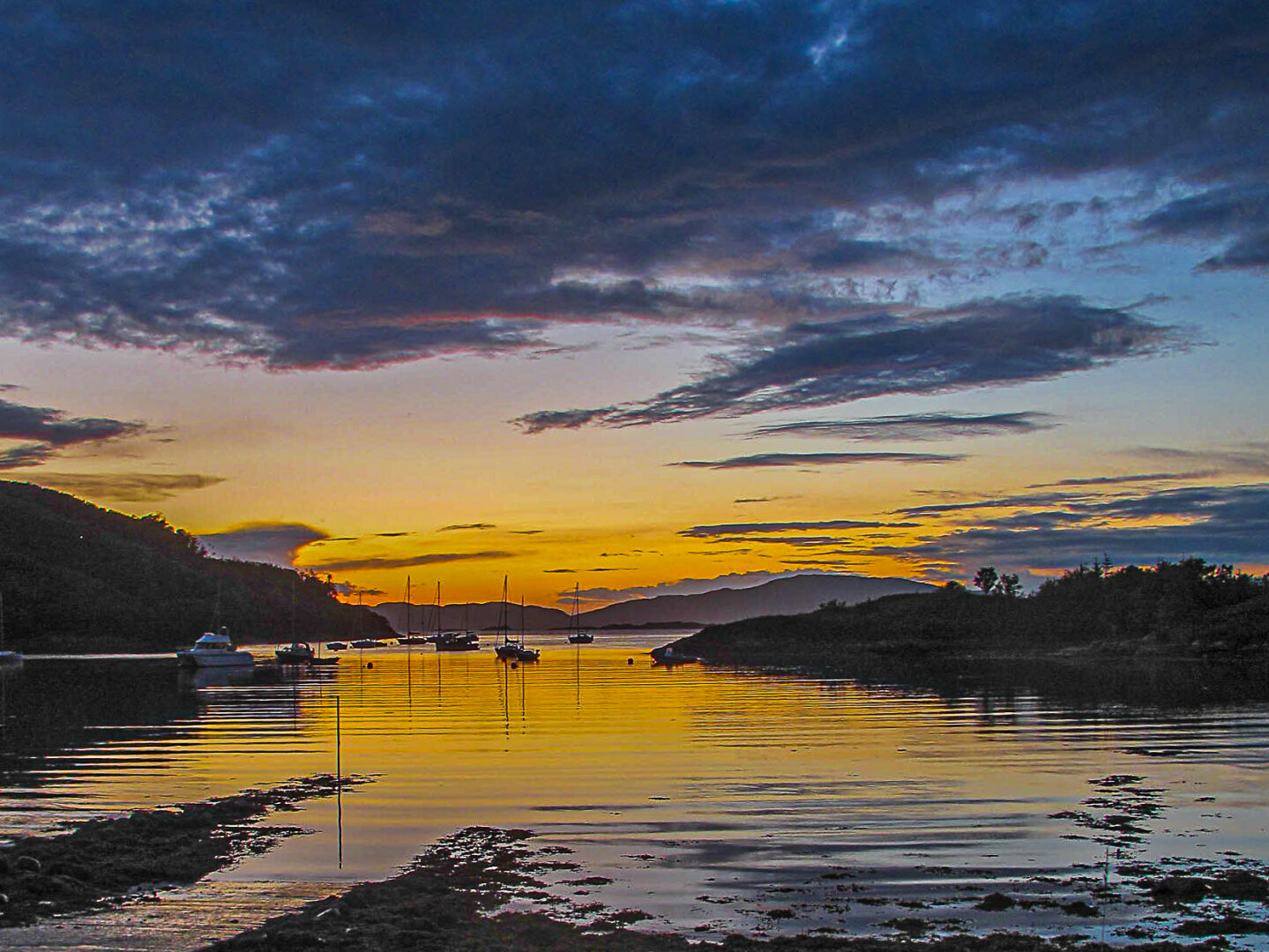 Golden sunset over Crinan Harbour in Argyll, Scotland, with moored boats reflecting on calm water. Dark hills and scattered clouds frame the glowing horizon, where orange and blue tones blend across the sky.