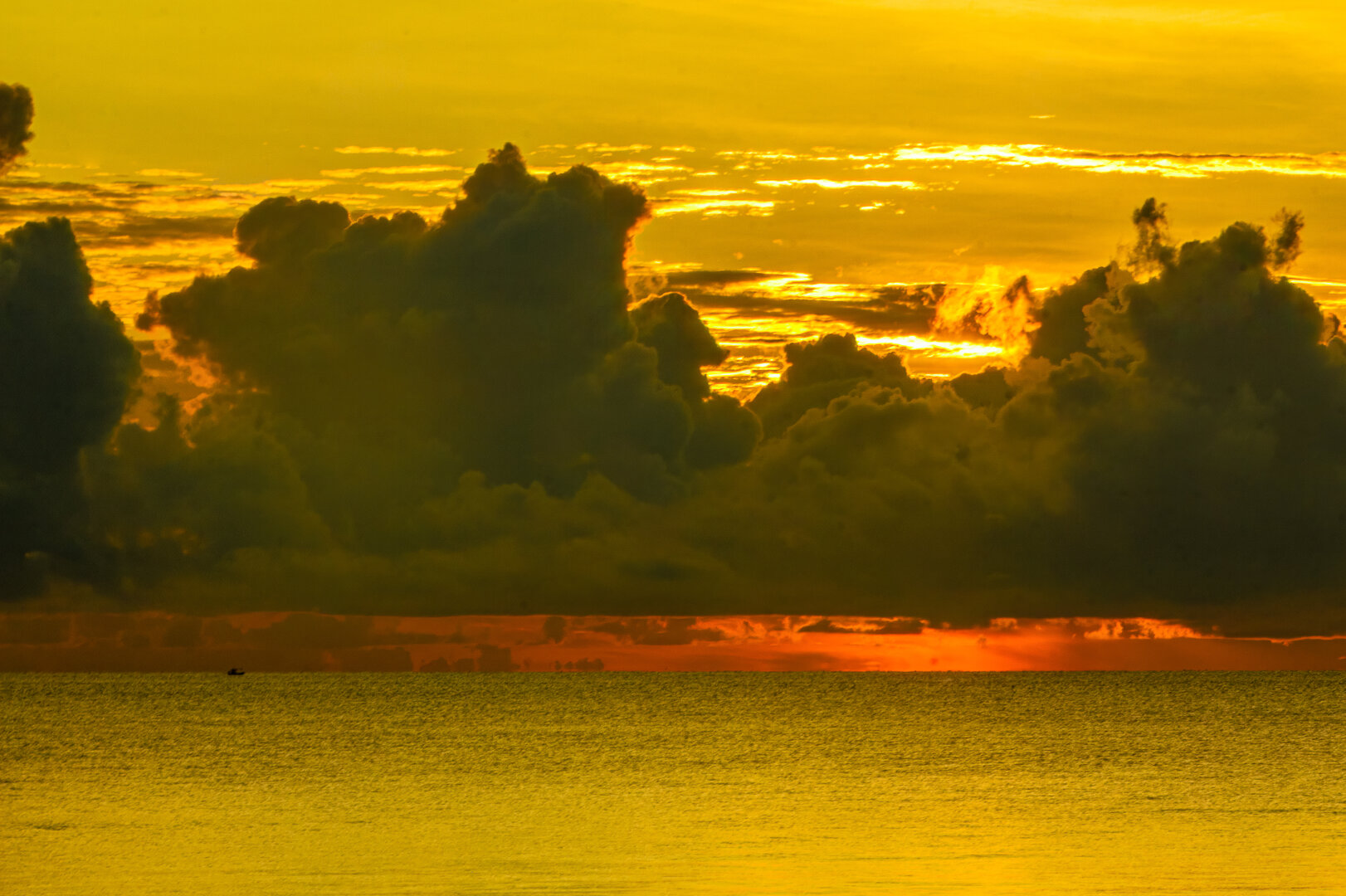 Golden dawn light illuminates towering monsoon clouds over the Gulf of Thailand. Dark, billowing storm clouds rise above a calm sea, while bands of bright yellow and orange sunlight break through gaps in the cloud cover near the horizon. A small boat appears as a silhouette against the glowing water, emphasising the dramatic scale of the tropical sky.
