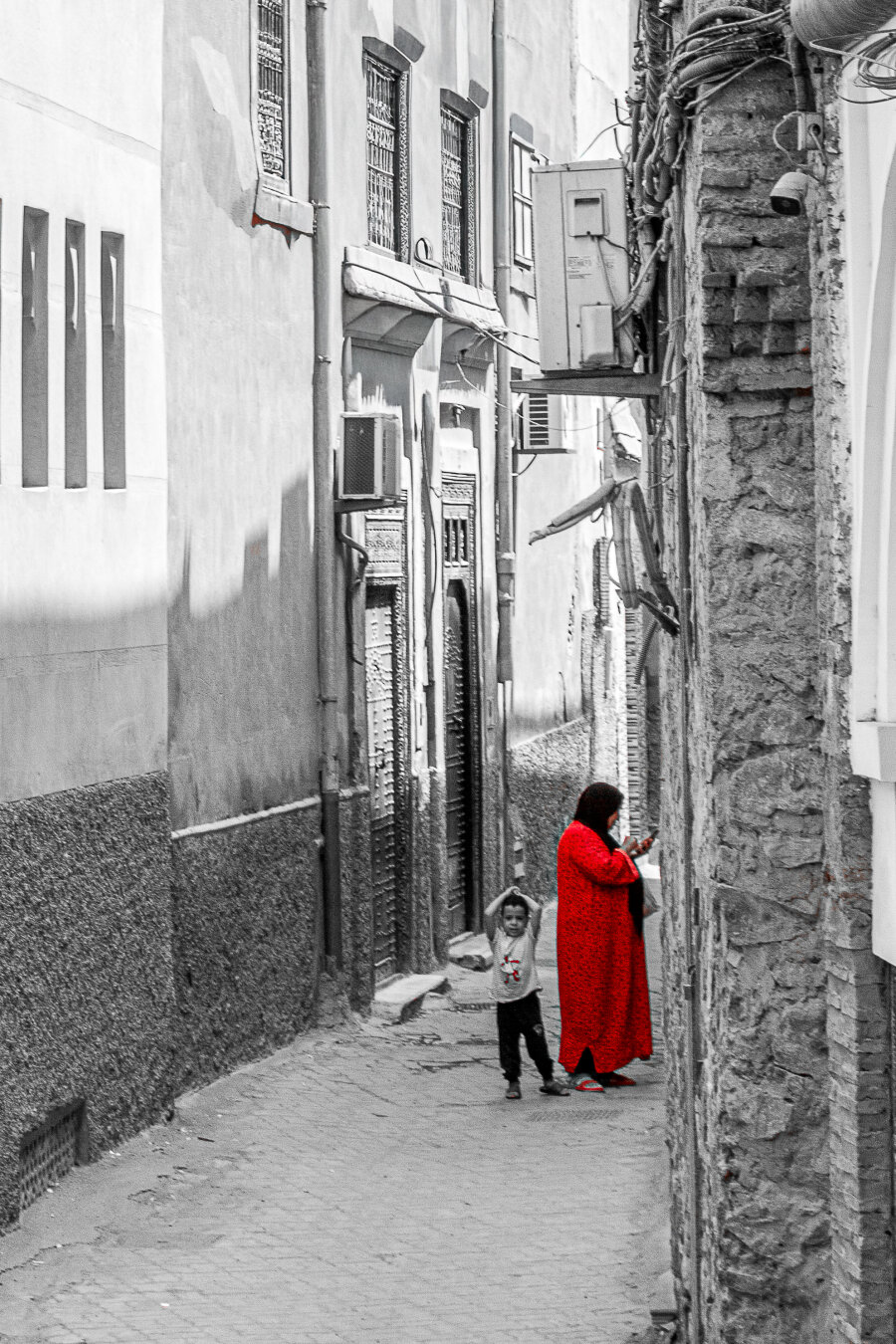 treet photograph taken in Marrakech, Morocco, showing a narrow cobbled alley lined with tall buildings and rough stone walls. The image is predominantly black and white, with selective colour highlighting a bright red cloak worn by a person standing near a child. The alley recedes into the distance, drawing attention to the contrast between the red garment and the monochrome urban setting.