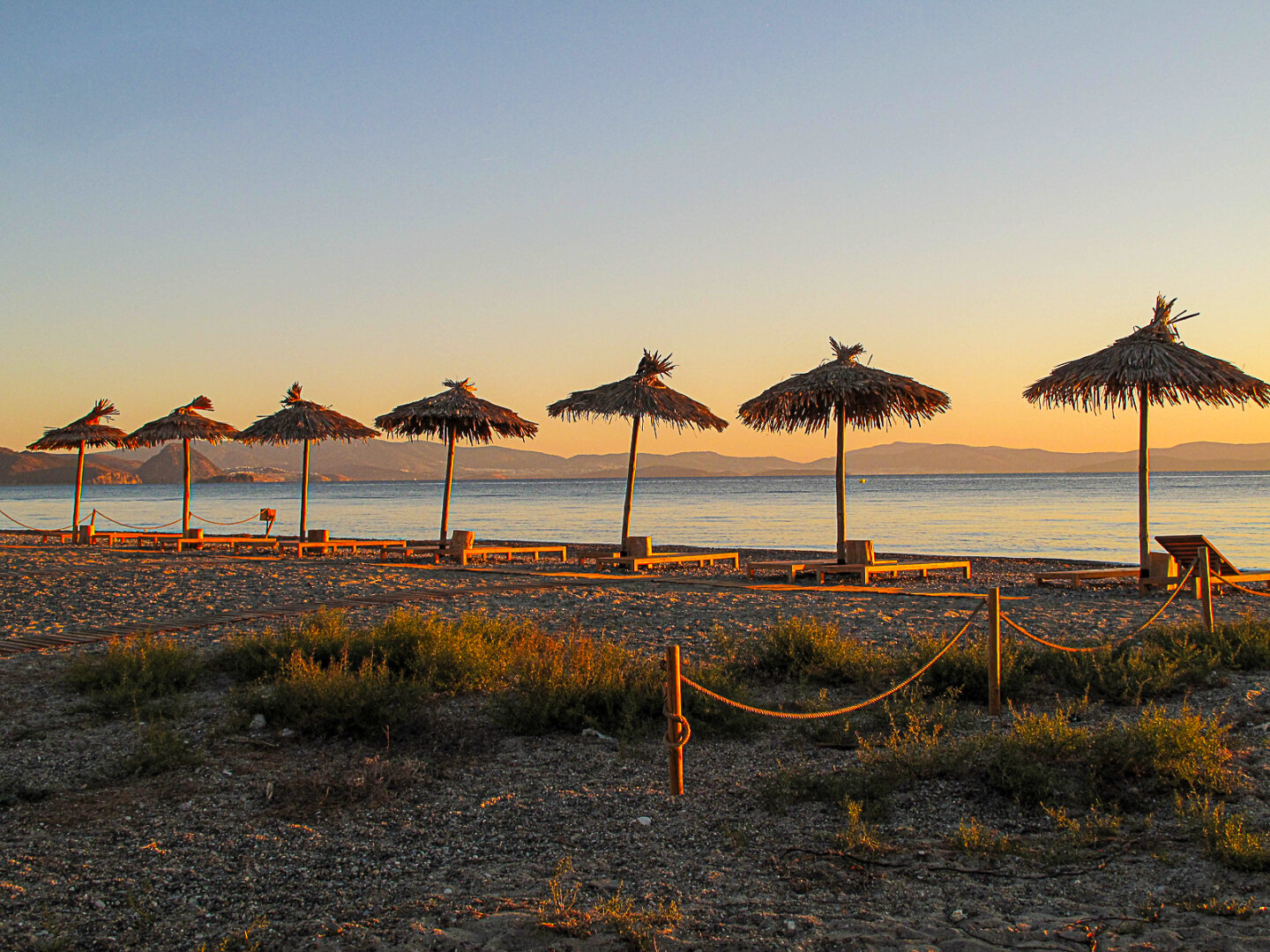 Early morning sunlight catches a row of thatched parasols and empty loungers along Lambi Beach, Kos, with calm blue water and layered hills on the horizon