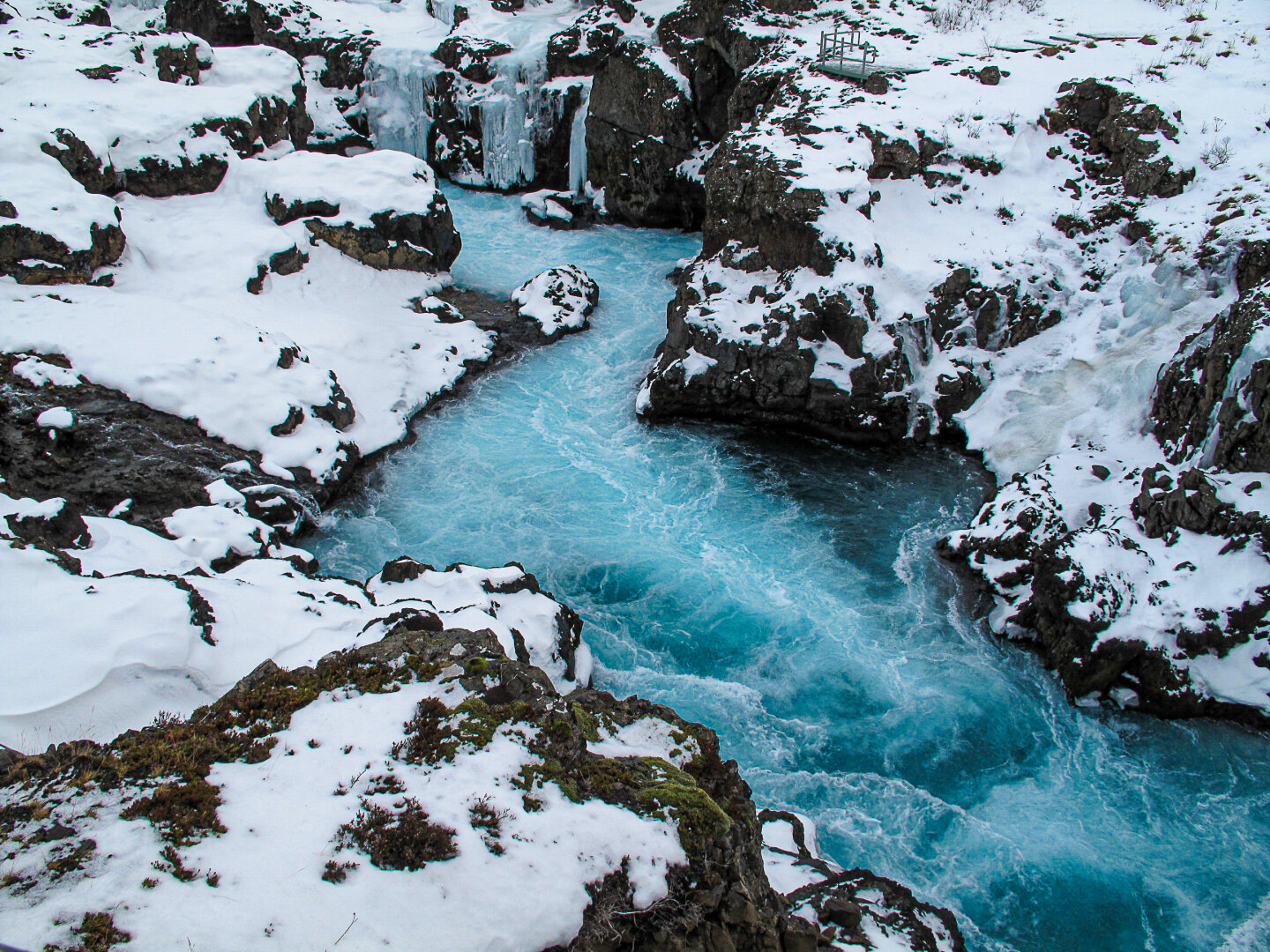 winter view of Barnafoss in western Iceland shows striking blue glacial water flowing swiftly between dark volcanic rocks blanketed with snow. Small frozen waterfalls and icicles cling to the cliff faces, while the river churns through the narrow canyon below. The contrast between the bright turquoise water, white snow, and black lava rock creates a dramatic Arctic landscape.