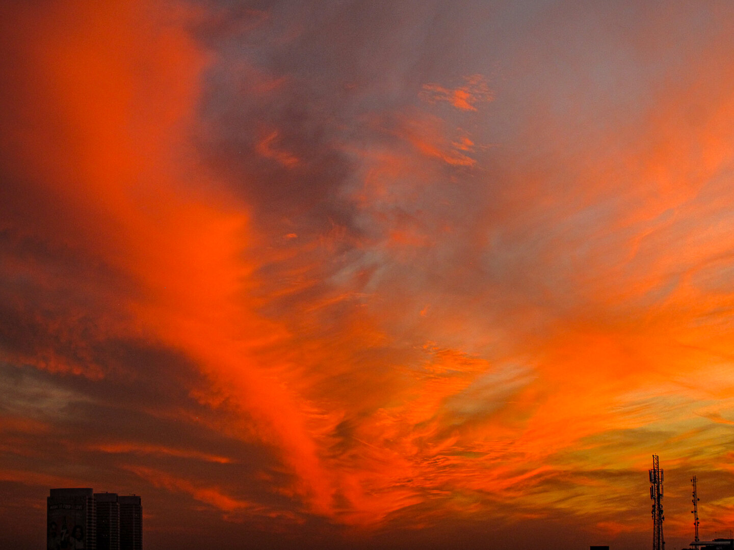 Fiery orange and red clouds stretch across the sky over Peshawar, Pakistan at sunset, with silhouettes of tall buildings and communication towers below.