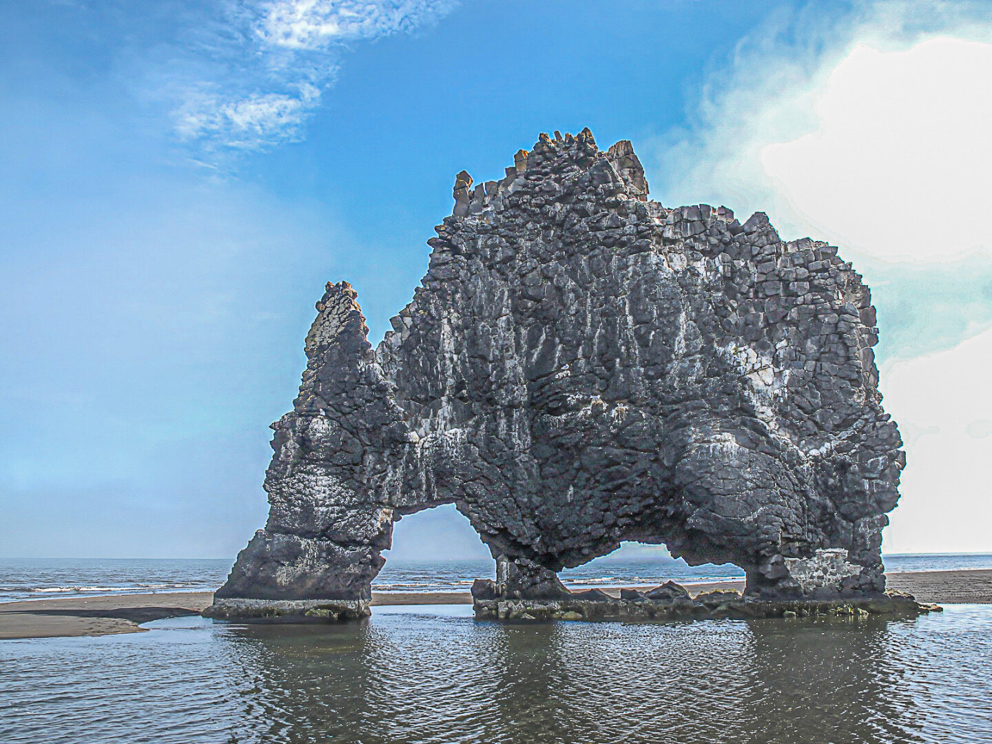 A large basalt sea stack known as Hvitserkur dominates the frame, standing in shallow coastal water at low tide in Vatnsnes, Iceland. The rock formation resembles a massive animal bent down to drink, with two natural arches forming its "legs." Dark volcanic stone, streaked with white bird guano, contrasts with the bright sky. Sea fog drifts around the coastline and blends into the horizon, creating a soft, atmospheric backdrop. Reflections ripple across the calm water in the foreground, while patches of sand are visible around the base of the formation. The cool tones of blue, grey, and black emphasise the remote and dramatic character of the Icelandic shoreline.

Provide your feedback on BizChat