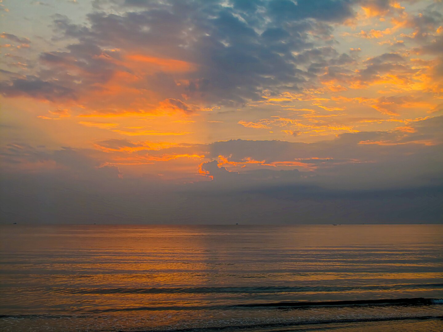 Dawn light spreads across the Gulf of Thailand at Pranburi, viewed from Aleenta. Layers of orange and gold clouds glow above the calm sea, while soft reflections shimmer across the water and gentle waves roll onto the shoreline below.