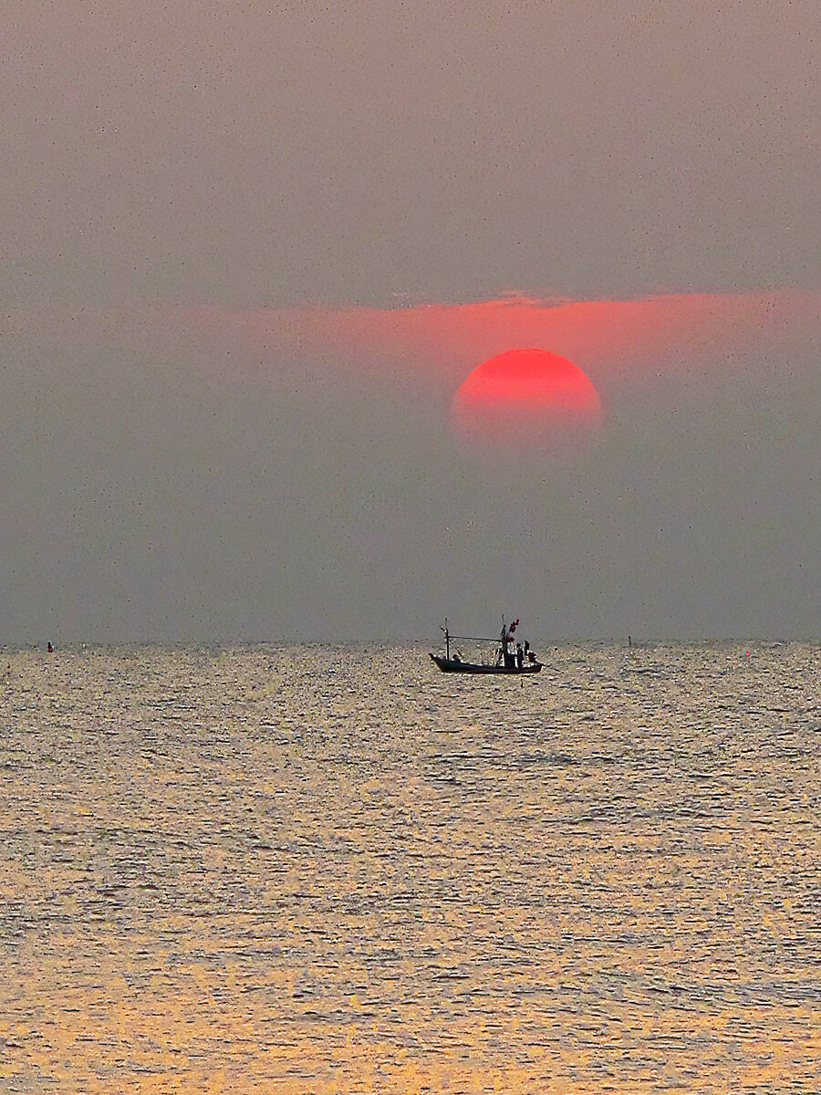 A small fishing boat carrying three people is silhouetted against the horizon off the coast of Pranburi, Thailand. A deep red sun rises through thin cloud and haze above the vessel, while soft golden light shimmers across the gently rippled water. The simple composition emphasises the contrast between the dark boat and the glowing sunrise, creating a peaceful early-morning coastal scene.