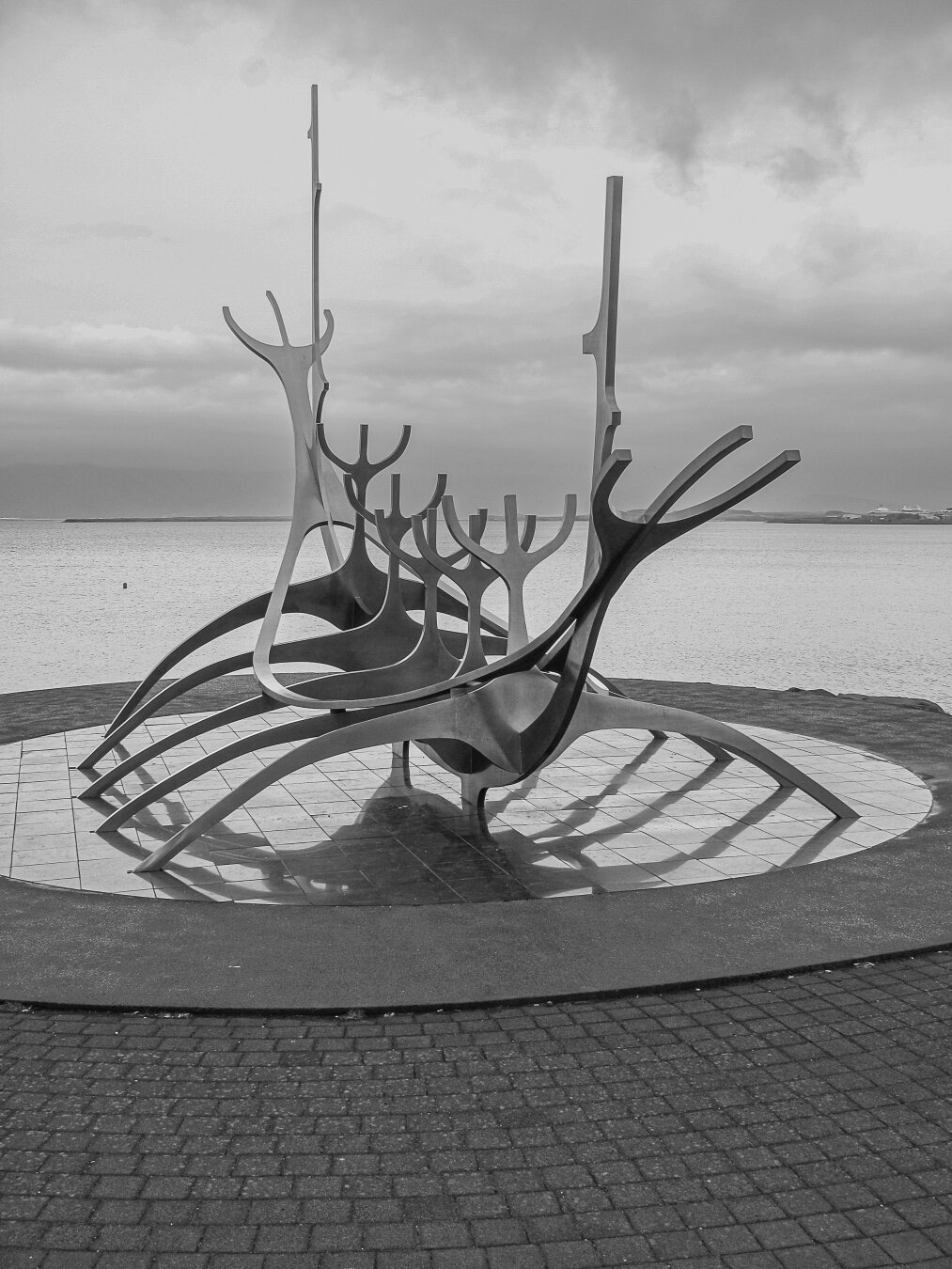 Black‑and‑white image of the Sun Voyager sculpture on Reykjavík’s waterfront. The polished steel structure, shaped like a stylized Viking ship with sweeping, upward‑reaching ribs, stands on a circular tiled base beside calm sea water under a cloudy sky.