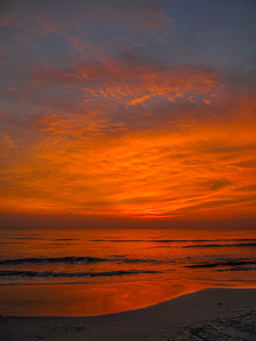 Dawn breaking over the Gulf of Thailand at Pranburi, viewed from Aleenta Resort. Brilliant orange and gold clouds fill the pre-sunrise sky above a calm sea, reflecting warm colours across gentle waves and wet sand along the shoreline.