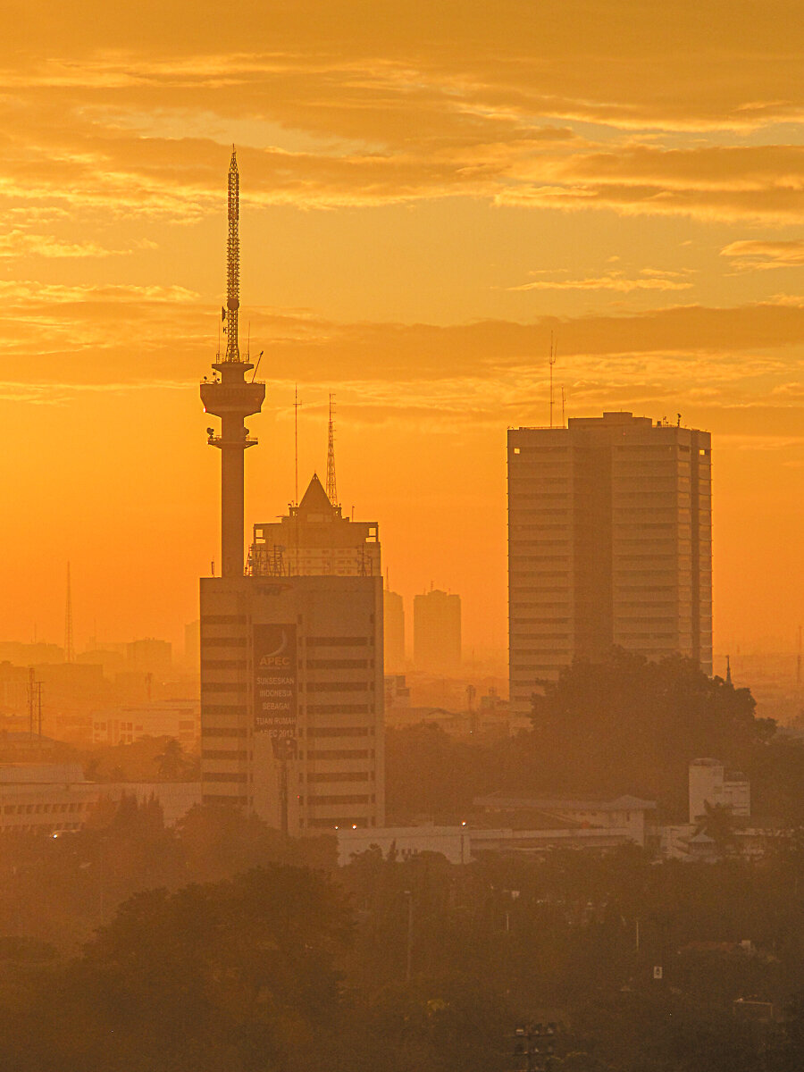 Early morning view of Jakarta, Indonesia, with high-rise buildings and a communications tower rising through a thick layer of orange-tinted smog. Warm sunrise light fills the sky and softens the distant skyline, while the lower cityscape fades into the haze.