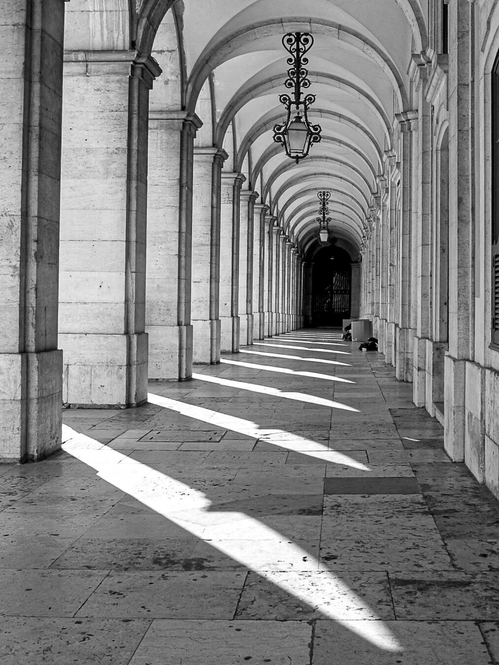 Black‑and‑white view of a long stone arcade in Lisbon, Portugal, with repeating arches and hanging lanterns; angled sunlight casts sharp triangular shadows across the tiled floor,