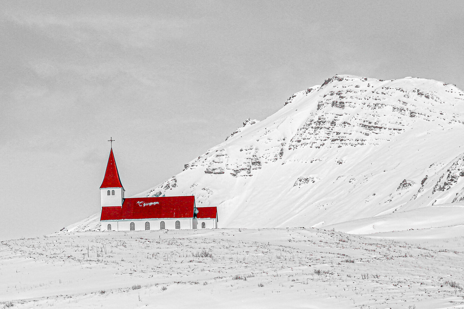 Hilltop church in Vik, Iceland, photographed in winter. The small white church and its bright red roof and steeple stand out against a largely black-and-white scene of snow-covered ground, a dramatic mountainside and a grey sky. The selective colour effect draws attention to the church, emphasising its isolation and contrast within the stark Icelandic landscape.