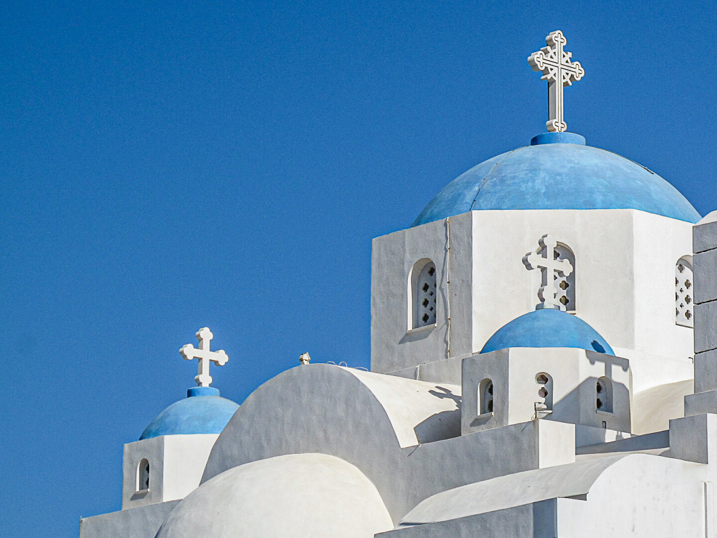 Photograph of a traditional Greek Orthodox church in Naxos City on the island of Naxos. The image focuses on the upper sections of the building, where whitewashed walls and curved roofs frame several blue-painted domes. Ornate white crosses crown the domes, and strong Mediterranean sunlight casts crisp shadows across the architecture. The rich blue sky fills the background, emphasising the striking contrast between the white buildings and the church's blue domes.