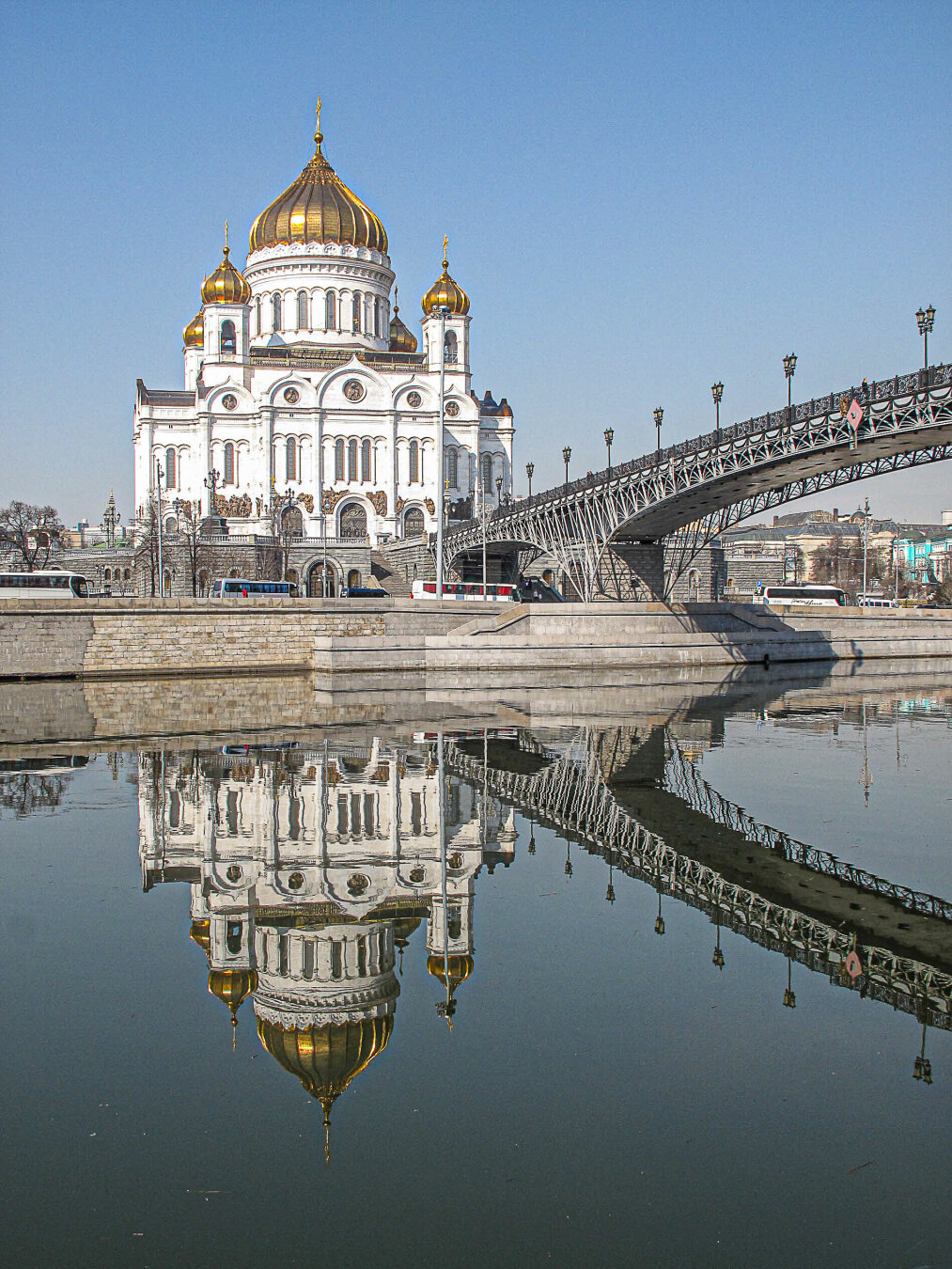 Cathedral of Christ the Saviour in Moscow rises behind a decorative steel bridge, its white façade and gold onion domes reflected almost perfectly in the still waters of the Moskva River. The calm river creates a mirror image of both the cathedral and bridge beneath a clear pale blue sky.