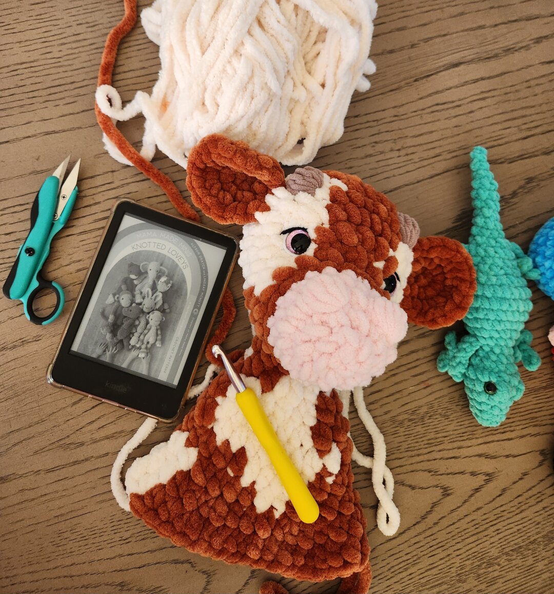 Partially completed crochet reddish brown and cream cow snuggly plush with a kindle, yellow crochet hook, and nippers