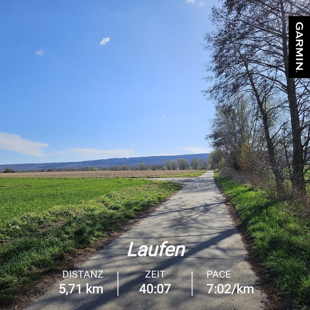 Blauer Himmel über dem Deister. Ein befestiger Feldweg mit Bäumen und bisschen auf einer Seite führt gerade auf den Deister zu. Die Sonne scheint, Schatten fallen auf dem Weg. Neben dem Weg ein schon grünes Feld. 
Laufdaten: Distanz 5,71km, Zeit 40:07, Pace 7:02/km