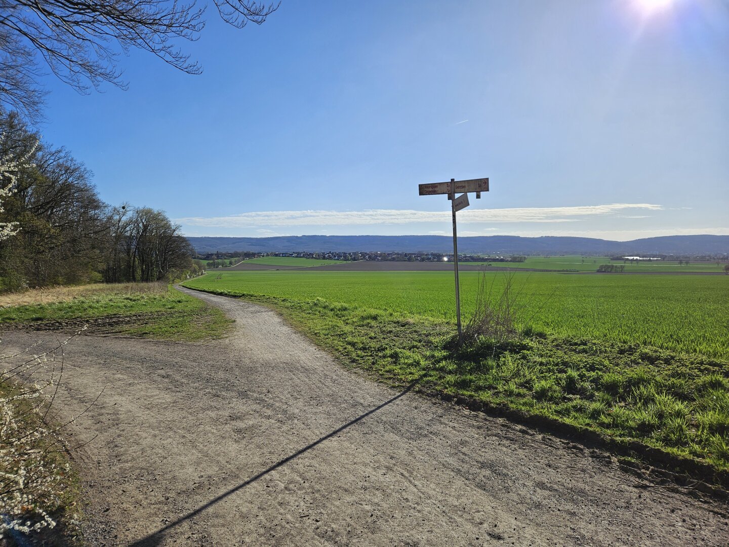 Blick vom Gehrdener Berg auf das Deister Vorland mit Degersen, Egestorf und Barsinghausen. Darüber blauer Himmel und Sonne. Im Vordergrund eine Feldweggabelung mit einem Wegweiser.