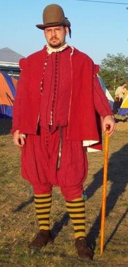 A white man wearing a a red linen Elizabethan suit of doublet and truckhose. Over that he wears a red wool cassock, topped off with a tall brown hat, striped knee socks, brown shoes and holds a walking stick.