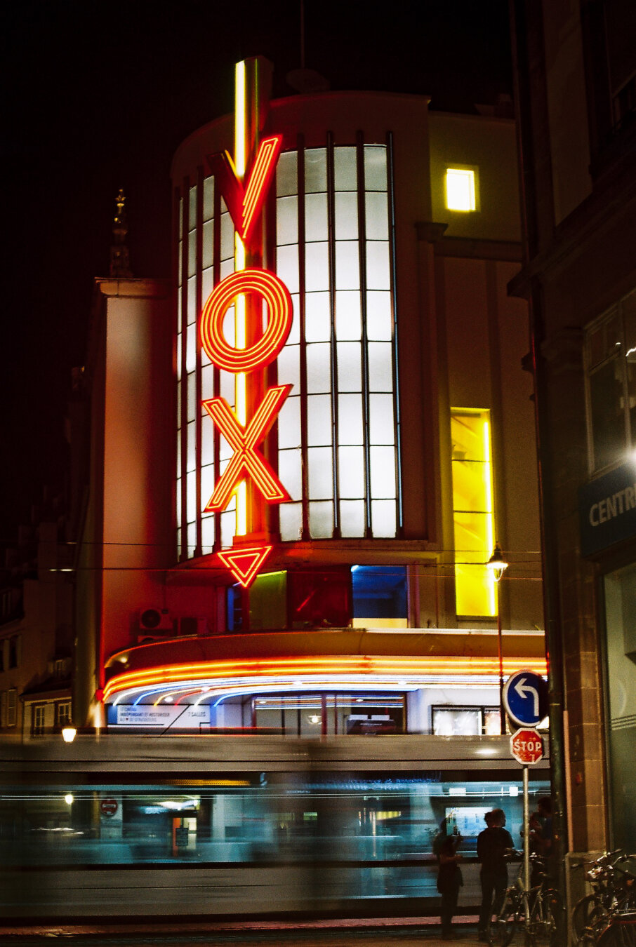 Nighttime long exposure photo of the Cinéma Vox in Strasbourg, with neon signage glowing brightly and light trails from a passing tram in the foreground. Taken on analog film with a tripod-mounted camera.