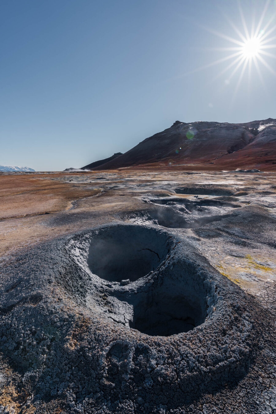 Wide-angle photo of a steaming mud pool at Námafjall Hverir with distant mountains in the background and a sunstar overhead on a clear, sunny day.