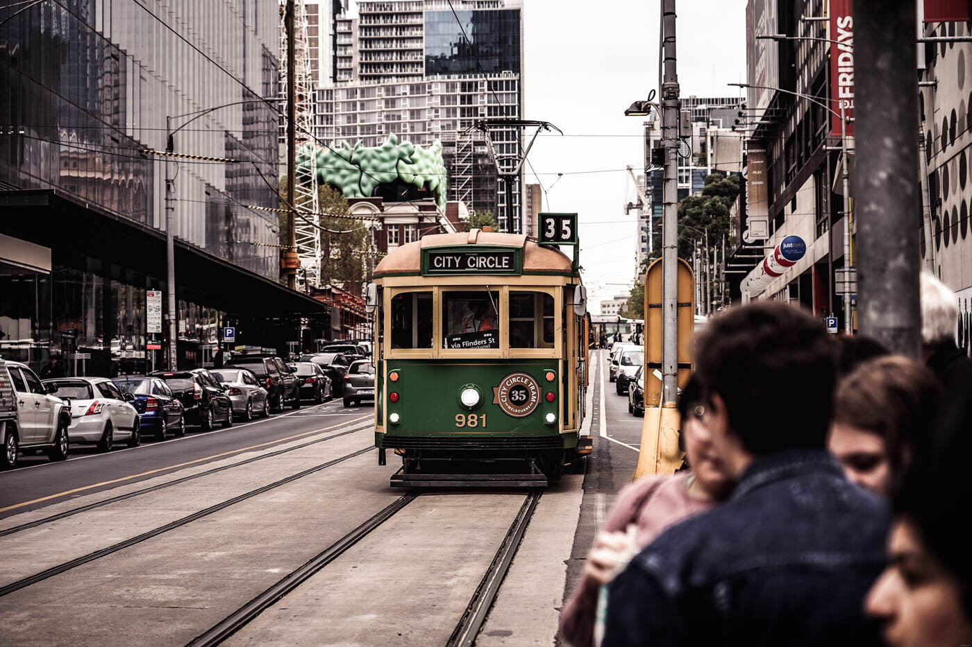 City Circle heritage tram approaching a stop in central Melbourne, with several people waiting in the foreground.