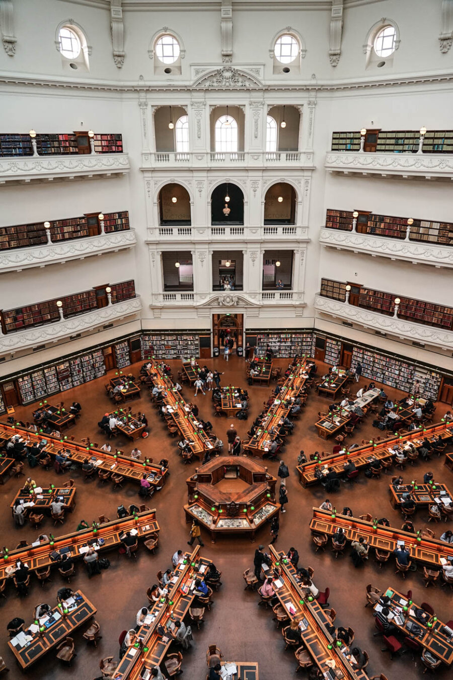 View from above inside the State Library of Victoria, showing the iconic reading room with its long wooden tables arranged in a star-like pattern and people quietly studying.