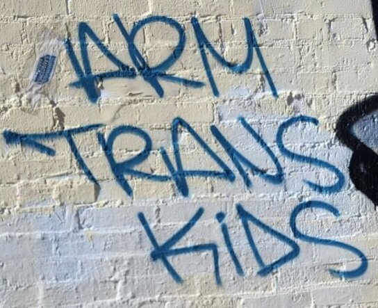 A dirty white painted brick wall with the words "ARM TRANS KIDS" spray painted in blue.