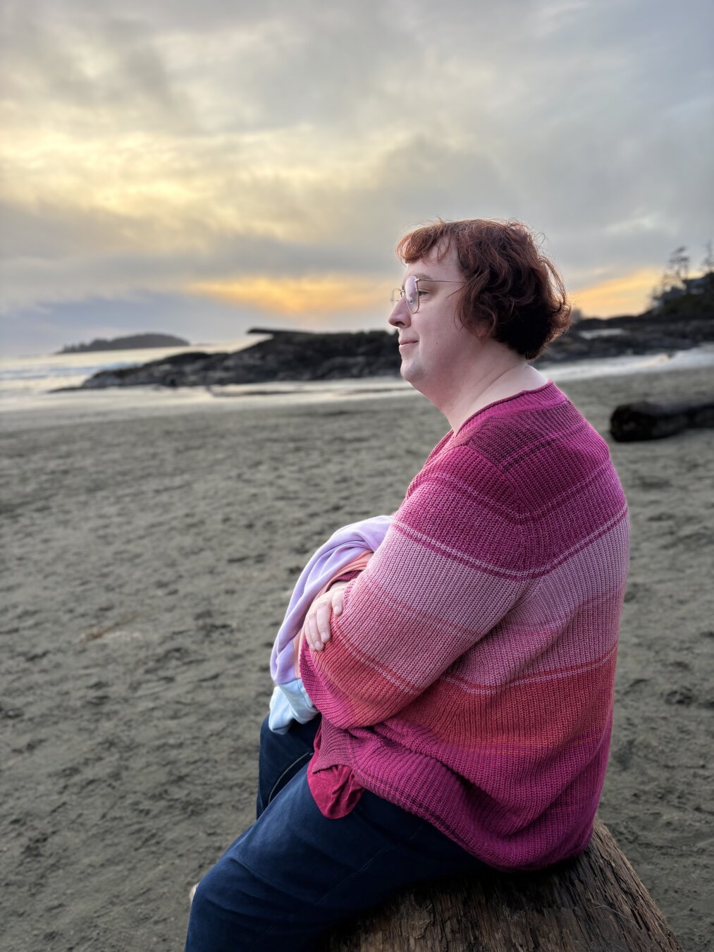 Sitting on a driftwood log while I look out to the sea. A sunset is behind me. I’m wearing a red/pink/stripped sweater and blue jeans, holding a hoodie in my arms. You can see my red hair and my glasses.