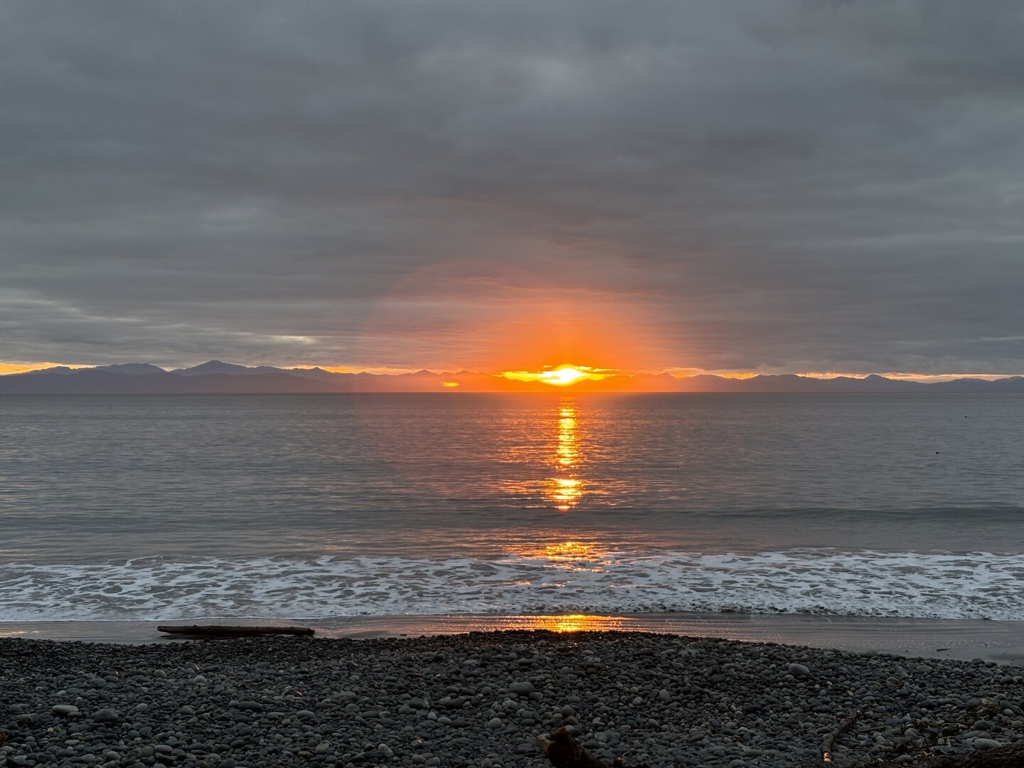 Looking out across the ocean towards Washington State. Most of the sky is grey and clouded but there’s a sliver of sky on the horizon in which an orange sky is visible and a bright spot in the centre is the sun.