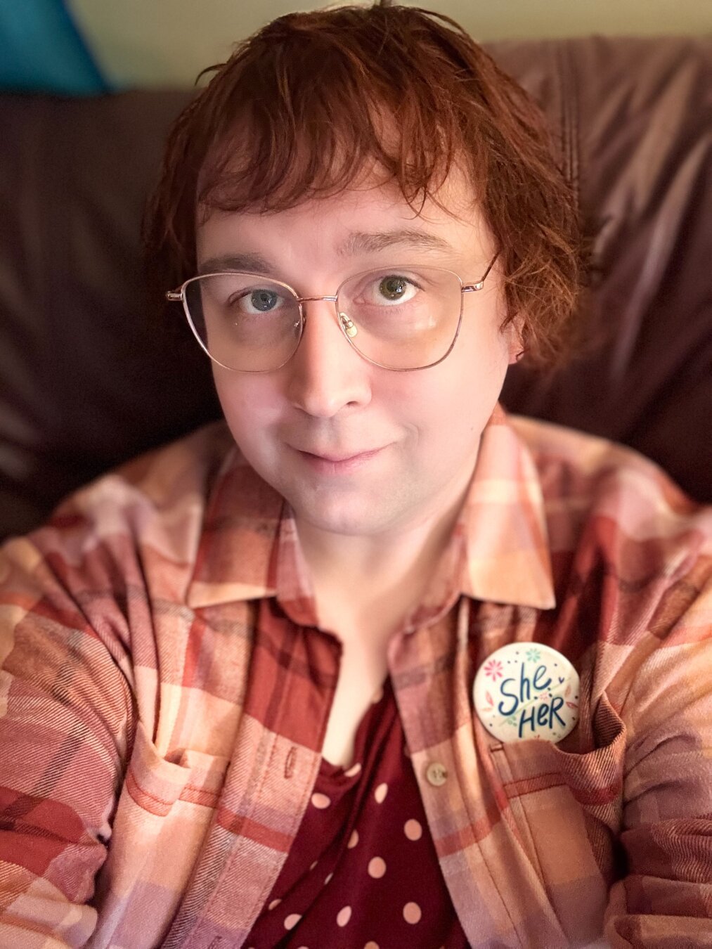 Lily posing for a selfie on her couch. She’s wearing a shirt with yellow spots on it, and a plaid shirt on top of that in the lesbian flag colours. She’s also got a She/Her pin on her left breast. Her reddish hair is curled up around the edges and hangs down in front of her face with bangs. She’s got rose gold glasses