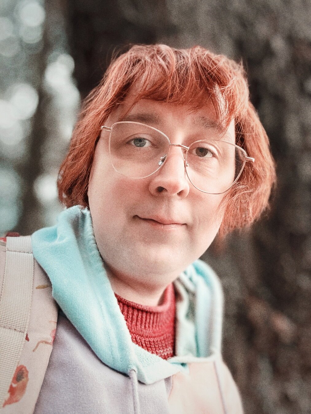 White trans girl looking at the camera with red hair in a bob with bangs and rose gold glasses, she has one blue eye and one brown eye, and she’s wearing a red sweater under a pink blue and purple pastel hoodie. The background is blurred but she’s in a forest next to the sea.