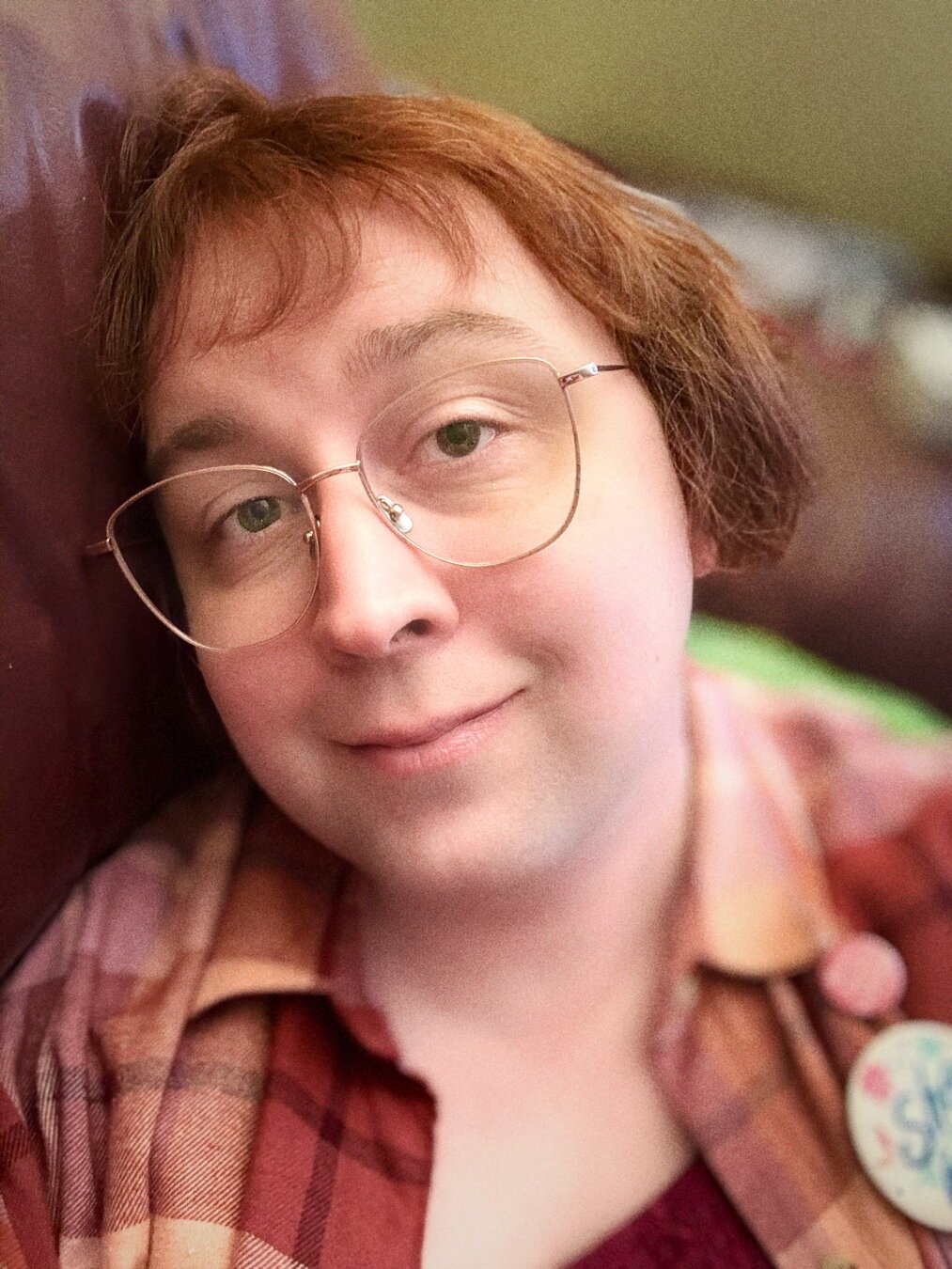 A white trans girl lying on her couch resting her head on a cushion, her reddish hair combed down around her ears and in bangs. Shes wearing cats eye glasses of rose gold, and a red/pink/orange plaid shirt with a She/Her button visible just off side. Shes smiling a little.