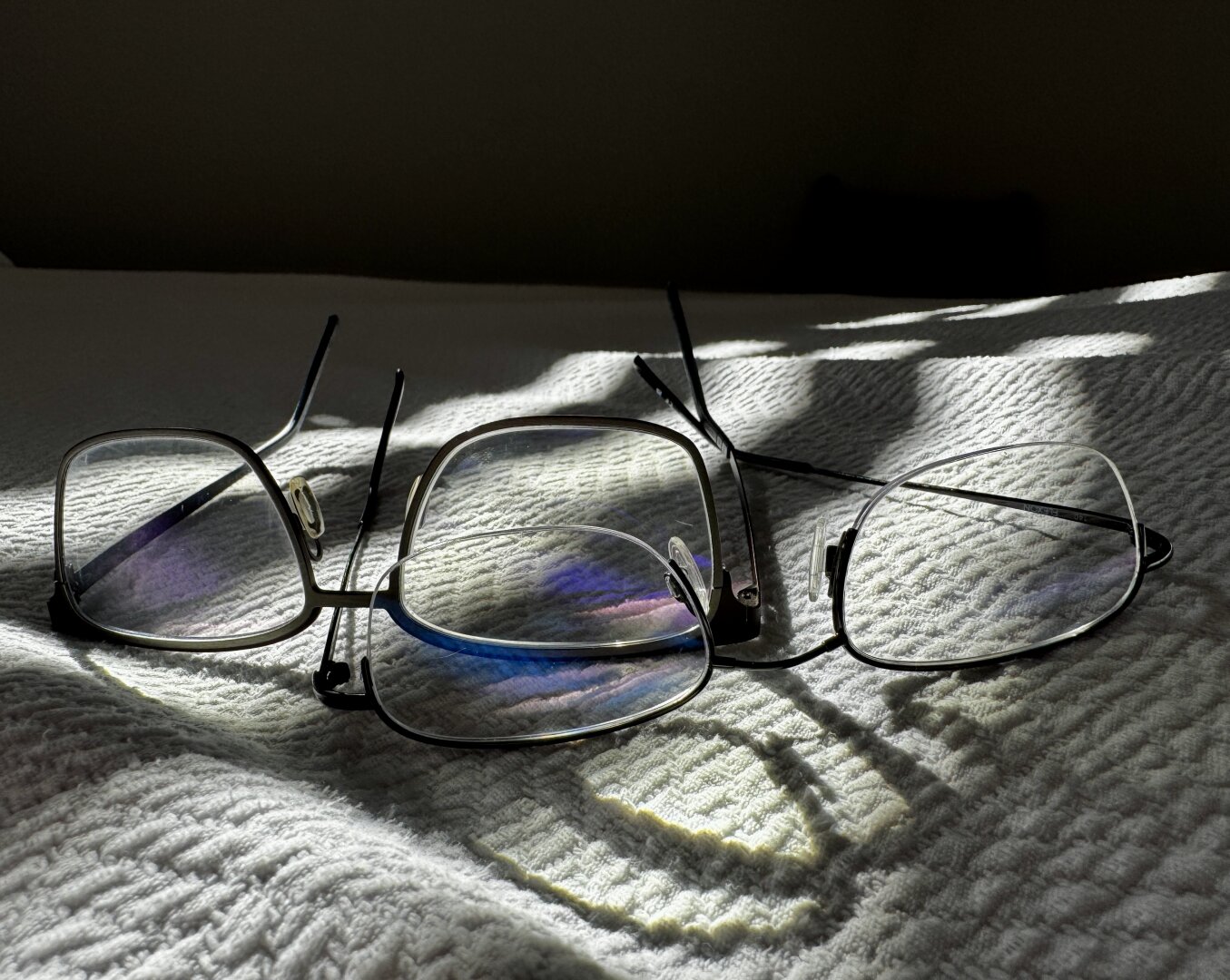 Two pair of eyeglasses, lying upside down on a white blanket, the sun streaking in through a nearby window
