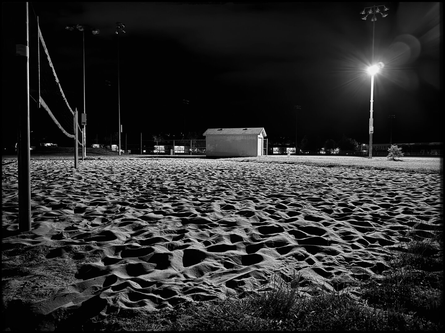 Black and white photograph of the University of Oregon’s sand volleyball courts. The perspective is oblique and low, with artificial lighting casting shadows across the ripples in the sand from use. Toward the right side of the frame stands a streetlight, which is the only source of light against a dark, night sky.