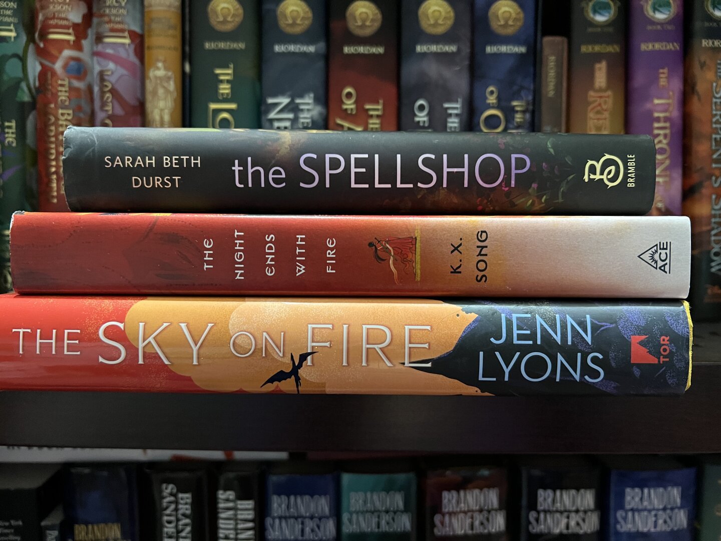 The spines of 3 books laying horizontally in front of other book spines. The titles visible are The Spellshop, The Night Ends with Fire, and The Sky on Fire