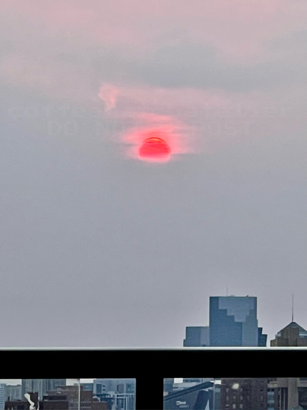 A red sun. The clouds surrounding the sun are red fading into a cloudy gray sky. The Minneapolis skyline at the bottom of the photo is slightly obscured by the metal rail and glass panes of the apartment building’a rooftop view.