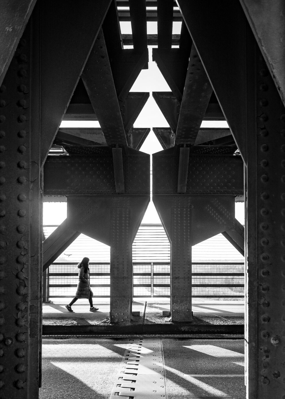 A woman walks on a bridge, where she is framed by steel i-beams and silhouetted by the setting sun.  In black and white.