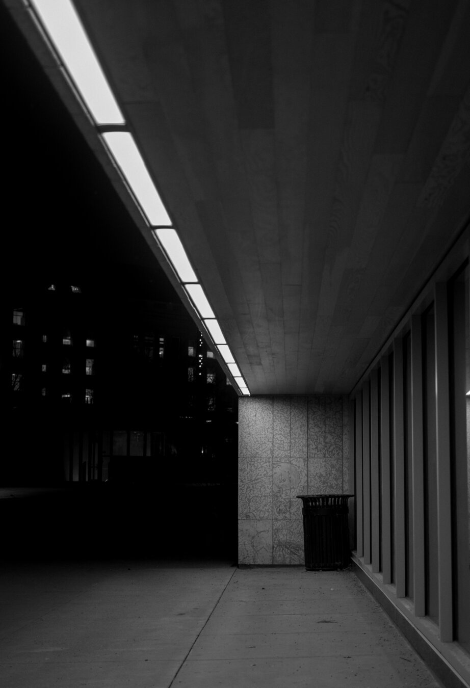 Strong brutalist architecture creates leading lines to a garbage can. Black and white photo.