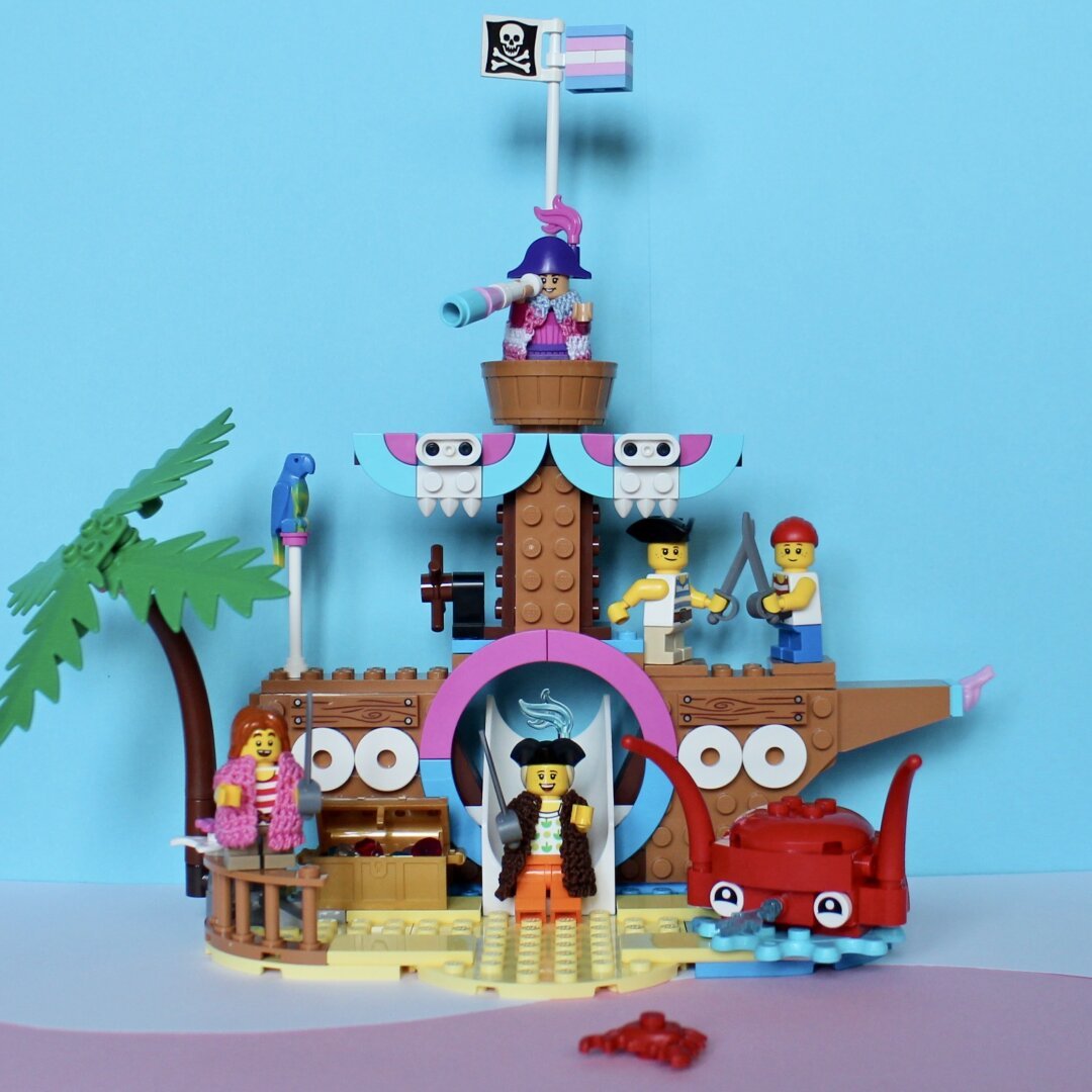 Lego pirate minifigs playing on a playground pirateship decorated with blue, pink and white parts.
Details: palm tree, crab, parrot, a pirate flag and a trans flag.