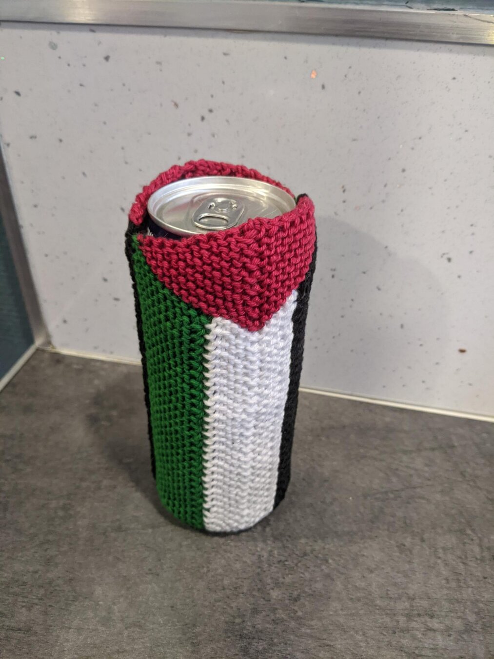 Photo of a tall drinks can wrapped up in a knitted Palestinian flag. The flag is position vertically with the red triangle at the top. The width of the flag is half the circumference of the can so there is also a second flag to make it a full cylinder but you can’t see it in the photo.