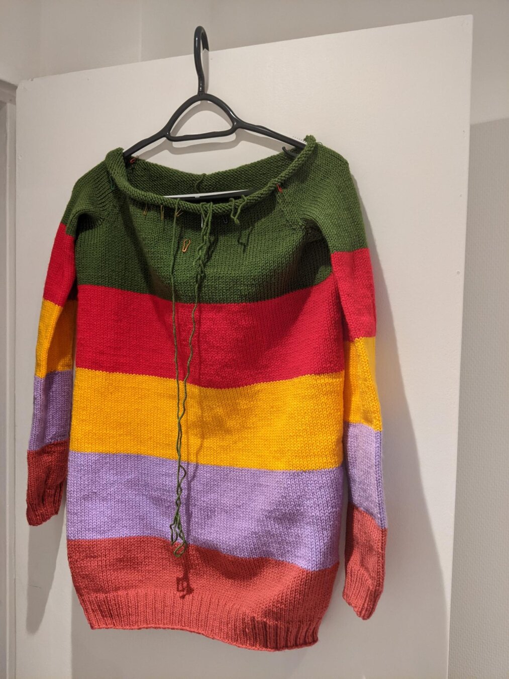 Un pull sur un cintre avec des rayures de couleurs verte, rouge, jaune, violette et rose. Le col du pull n’est pas terminé et il y a un long fil vert qui tombe.