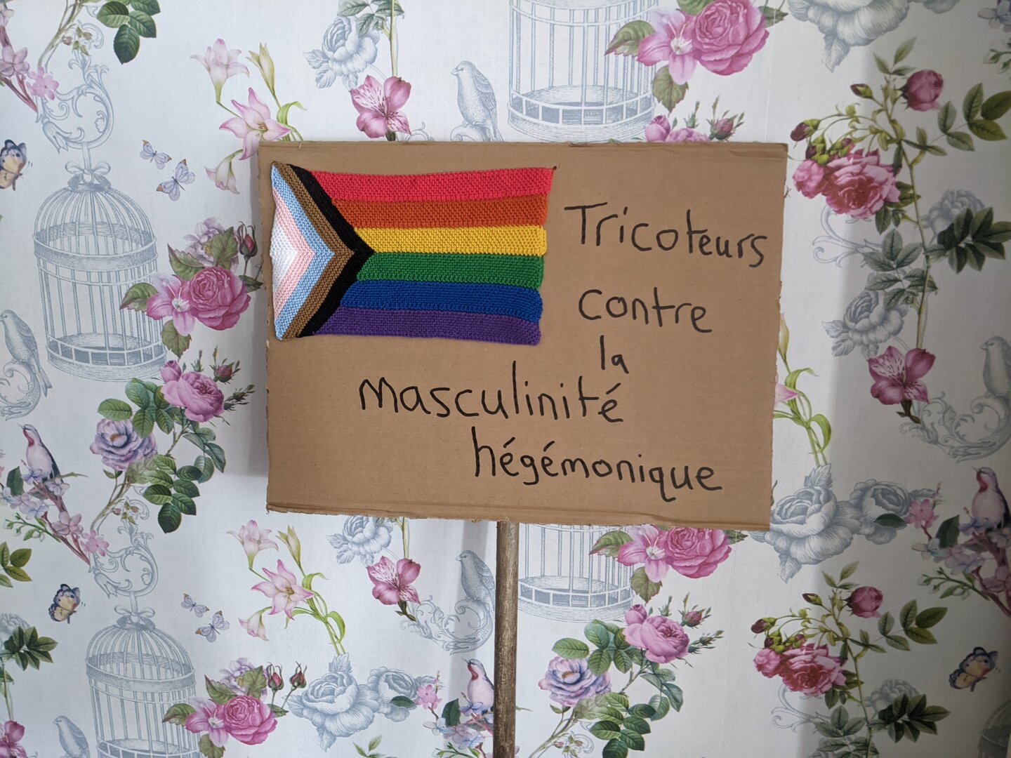 A cardboard protest sign attached to a wooden pole with the text "tricoteurs contre la masculinité hégémonique". A knitted progress pride flag is attached to the the top-right corner.