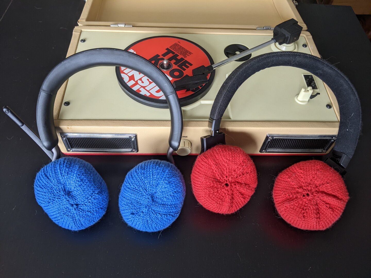 Photo of two pairs of headphones leaning against a record player for decoration. They’re the kind that go over your head. The left ones have a blue knitted cover over the ear part and the right ones have a red cover.