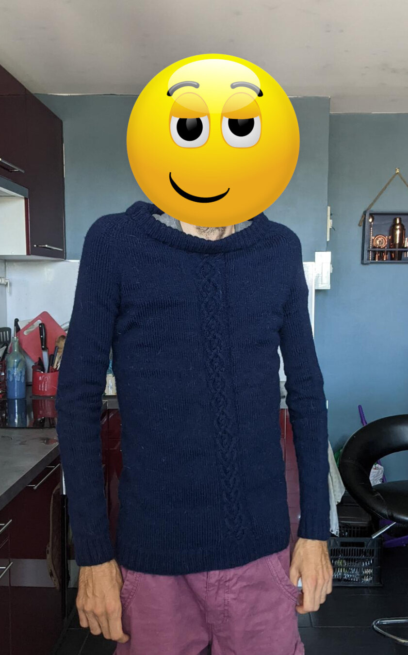 Photo of me wearing the jumper in my kitchen. My face is covered up with a smiley. The jumper is dark blue. It has a cable pattern running up the middle. The sleeves are quite tight but the collar is really loose.