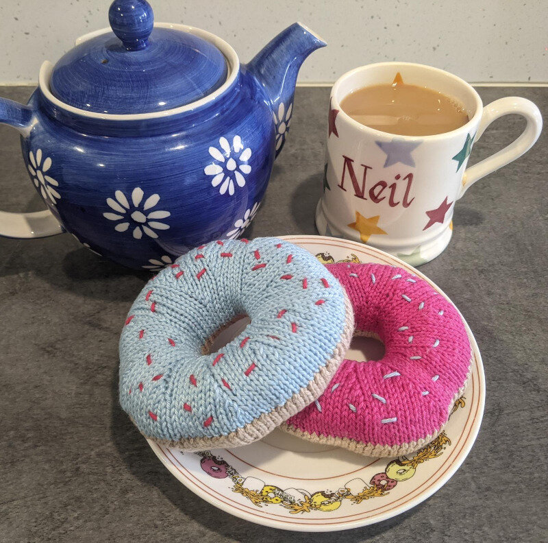 Two knitted doughnuts sitting on a plate. One has light blue icing and the other has pink. They both have a thread going through the top to look like sprinkles. The plate is Simpsons-themed and if you look closely there is a cup of coffee on it and something about dunking doughnuts. In the background there is a teapot and a cup of tea. The mug has my name on it (Neil).
