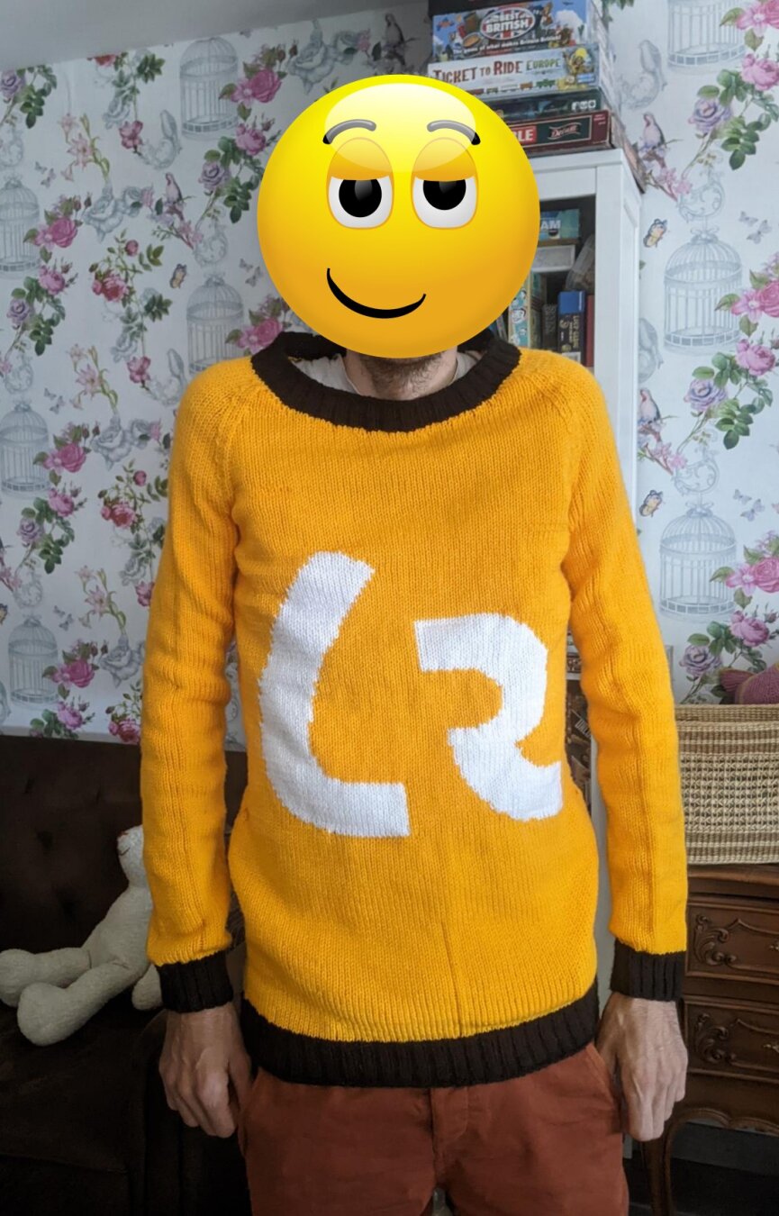 Photo of me wearing an orange/yellow knitted jumper. My face is covered by an emoji. The jumper has black cuffs, collar and rim at the bottom. On the front there are the large white letters 𐑖 and 𐑷 from the Shavian alphabet.
