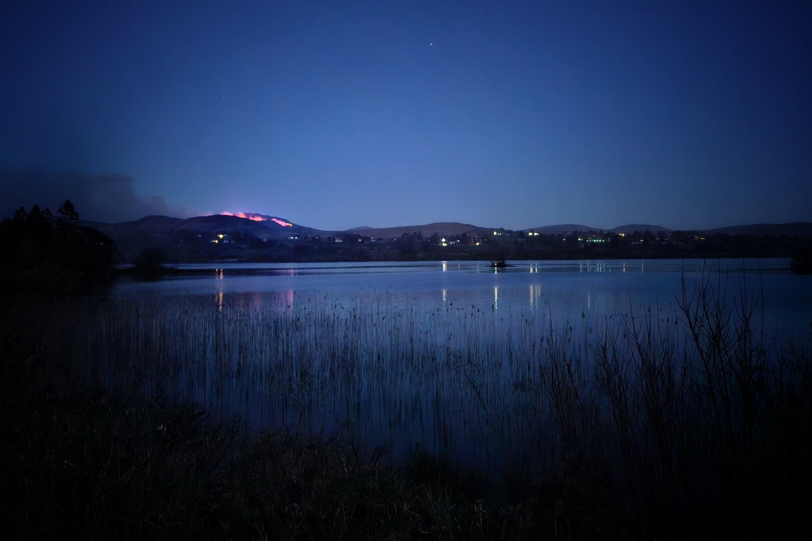 A wide shot of a calm lake at night, reflecting the dark blue sky. In the background, a hill is partially ablaze, casting a warm orange glow against the dark landscape.